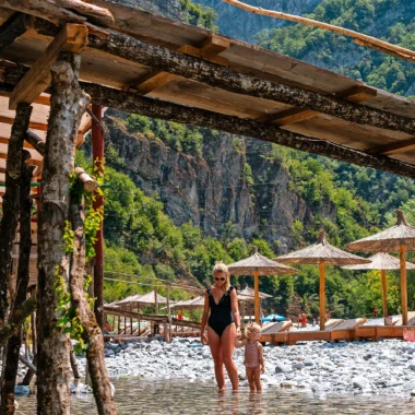 Moeder en kind lopen hand in hand door het ondiepe, kristalheldere water van de Shala-rivier in Albanië, omgeven door een spectaculair berglandschap en traditionele parasols op de achtergrond.