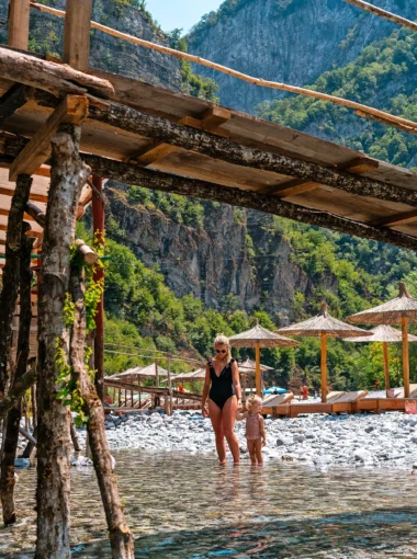 Moeder en kind lopen hand in hand door het ondiepe, kristalheldere water van de Shala-rivier in Albanië, omgeven door een spectaculair berglandschap en traditionele parasols op de achtergrond.