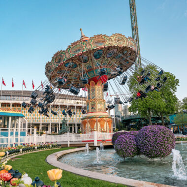 Decoratieve zweefmolen in Tivoli Gardens in Kopenhagen, omringd door kleurrijke bloemen, fonteinen en klassieke architectuur, met een blauwe lucht op de achtergrond.