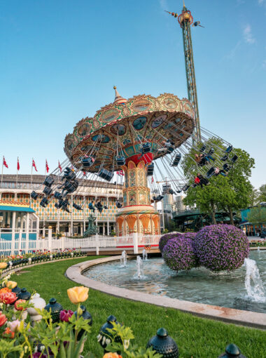 Decoratieve zweefmolen in Tivoli Gardens in Kopenhagen, omringd door kleurrijke bloemen, fonteinen en klassieke architectuur, met een blauwe lucht op de achtergrond.