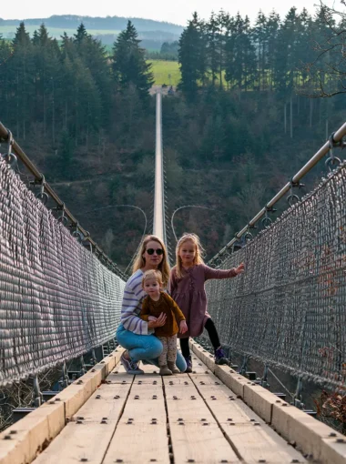 Moeder met twee jonge kinderen poseert op de Geierlay-hangbrug in Mörsdorf, Duitsland, met een bosrijke vallei en heuvelachtig landschap op de achtergrond.